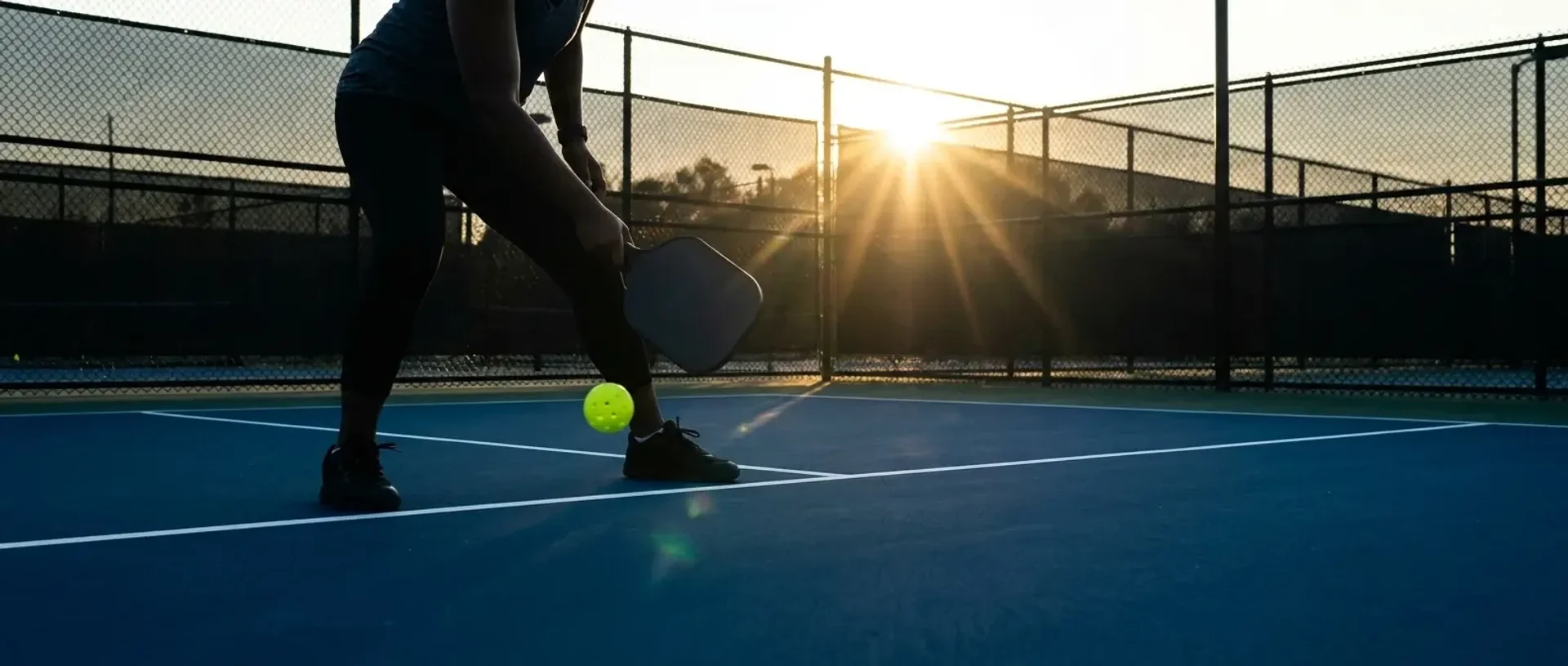 A pickleball server captured from the referee's perspective during a legal drop serve, both feet behind the baseline and paddle below the waist — illustrating the 2026 serve rules.