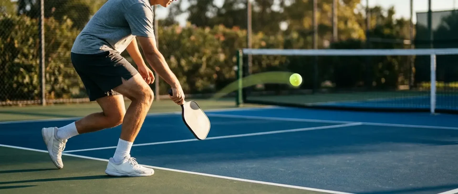 A pickleball player at the baseline watches a neon-yellow ball arc toward the opposing kitchen line — illustrating how to hit a third shot drop.